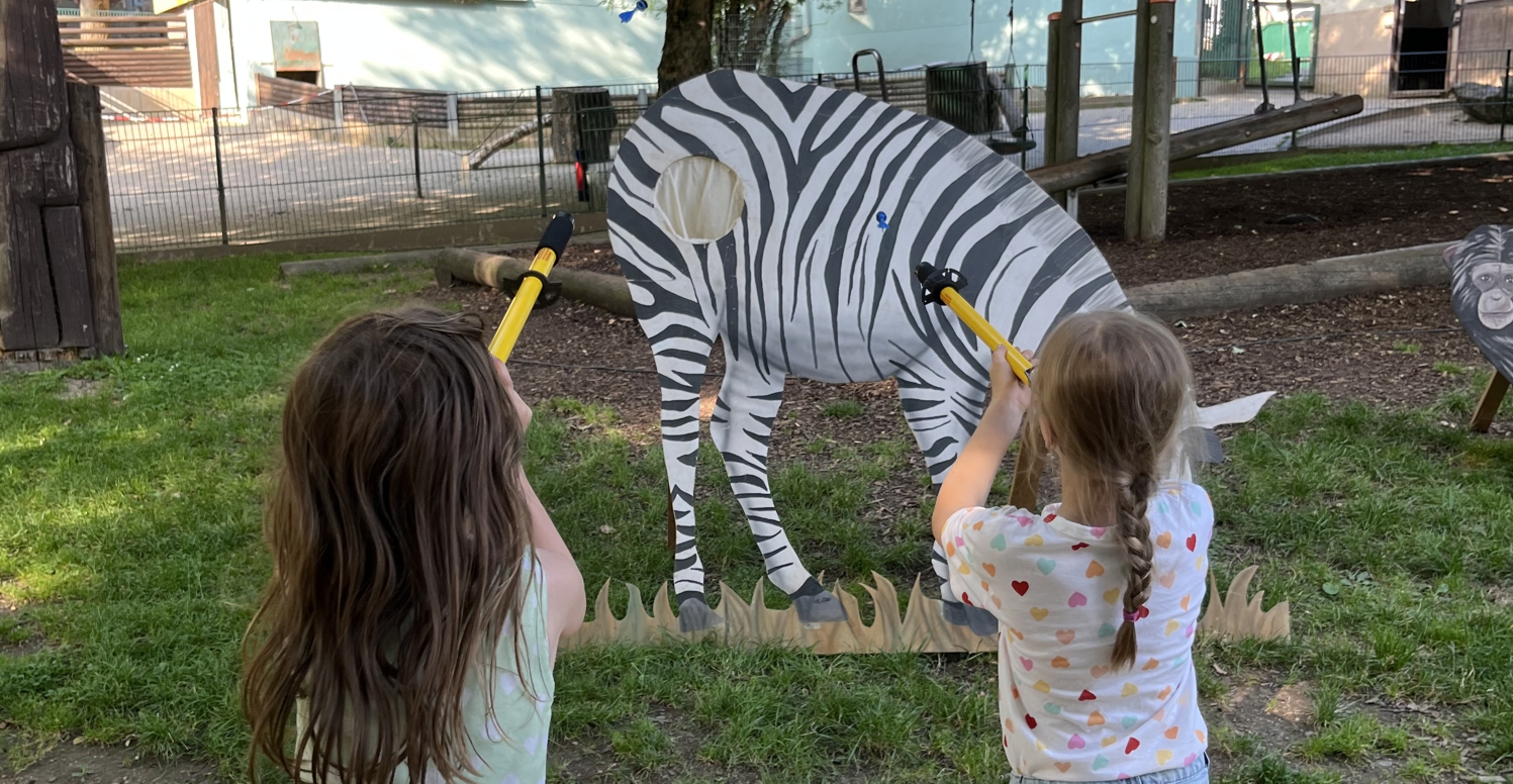 Zwei Mädchen pusten durch ein Plastikrohr Gummipfeile auf ein hölzernes Zebra